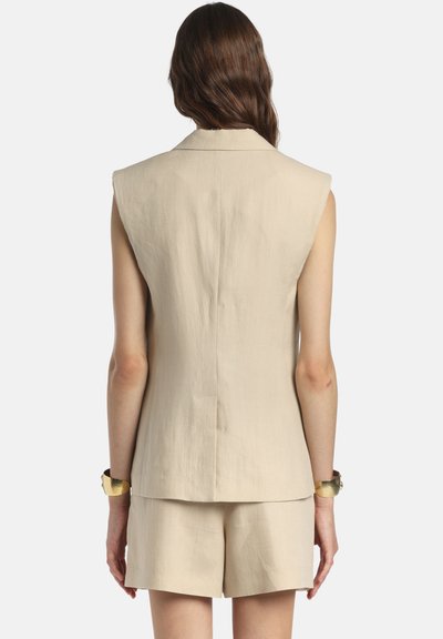 Femeie cu păr lung și castaniu poartă un sacou bej fără mâneci și pantaloni scurți asortați, văzută din spate, având un fundal simplu.