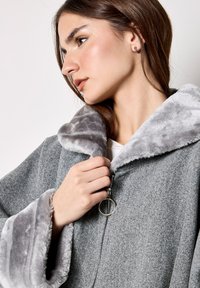 Cappotto grigio con zip, collo e polsini in pelliccia sintetica grigia soffice. Presenta un cursore della zip rotondo in metallo e un tessuto dalla trama evidente.