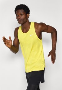 Nike gul topp med ermer som er gult, har et strukturert mønster, lett stoff og en liten hvit logo. Bært med svarte shorts.