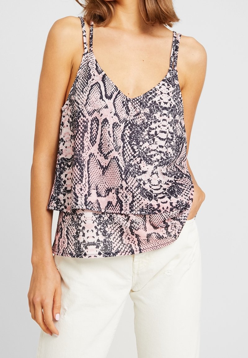 Femme portant un top caraco sans manches superposé rose et noir imprimé serpent avec de fines bretelles et un pantalon blanc, main dans la poche.