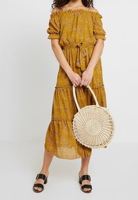 Off-shoulder klänning i senapsgul med blommönster, volangärmar och lager på lager i kjolen. Matchas med en rund vävd väska och svarta sandaler.