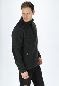 Maglione in pile grigio scuro con colletto alto, zip frontale, dettagli neri sulle spalle e tasca laterale con zip. Finitura in tessuto testurizzato.