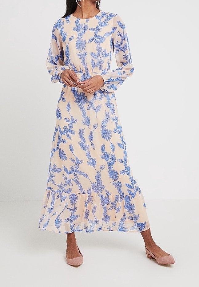 Femme portant une robe beige à manches longues, longueur cheville, à motif floral bleu et ourlet à volants, associée à des chaussures en daim roses à talons bas.