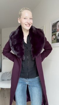 Cappotto lungo di un profondo colore viola con colletto in pelliccia sintetica, dotato di tasche laterali. Indossato sopra una maglia nera e jeans blu, esposto su uno sfondo neutro.