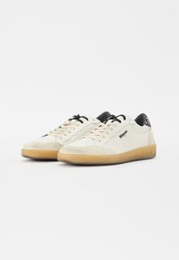 Hvite semskede og skinn sneakers med svarte detaljer, perforert tåparti og såle av gummi. Snøring og logo er synlig på siden.
