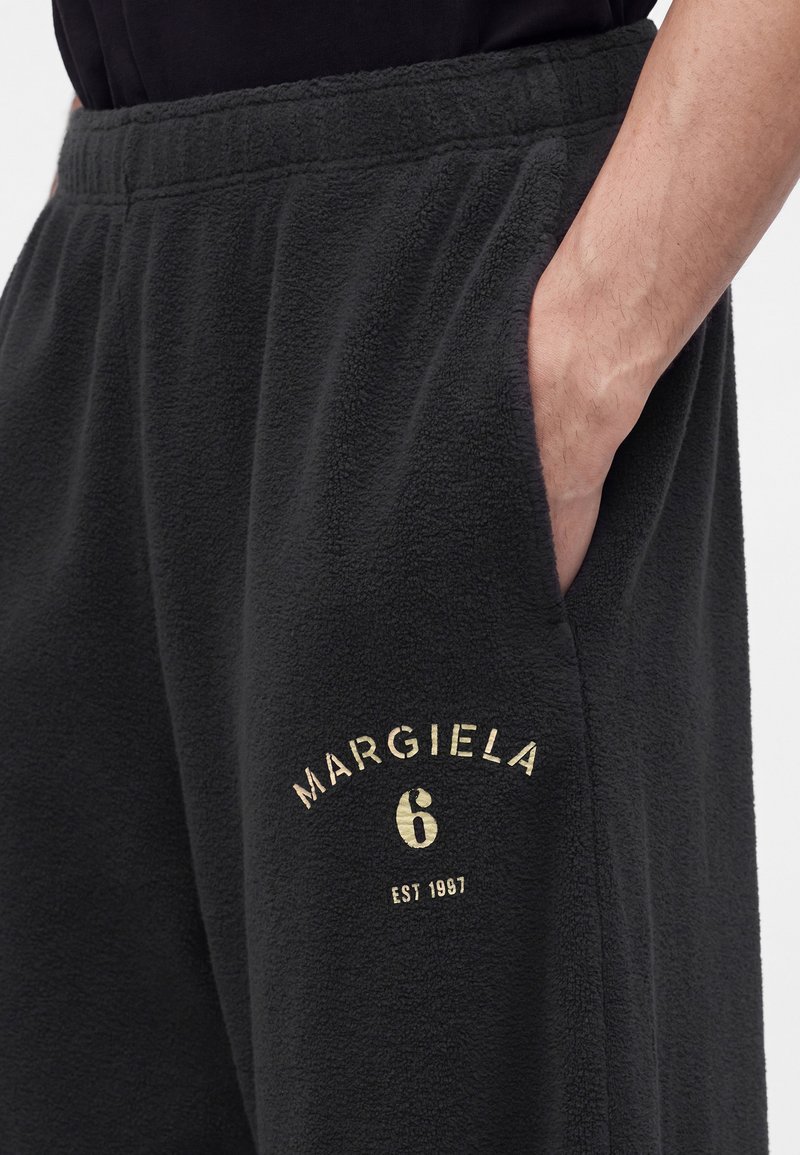 Nahaufnahme von schwarzen Fleecehosen mit elastischem Bund, Hand in der Tasche und „"MARGIELA 6 EST 1997"“ beige auf dem Oberschenkel gedruckt.