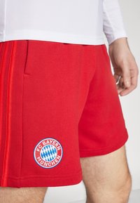 Bermudas deportivas rojas de poliéster, con un logotipo bordado del FC Bayern München y detalles en azul. Textura suave y corte atlético.