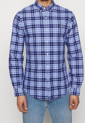 Homme portant une chemise à carreaux bleue, blanche et bleu marine à boutons, avec un jean bleu clair, debout devant un fond uni.