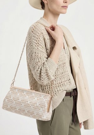 Vrouw die een beige opengewerkt trui en hoed draagt, houdt een beige handtas met patroon en een gouden kettingriem vast met een lichte jas over haar schouder.