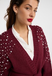 Cardigan burgundiu cu accesorii din perle albe pe mânici, textură tricotată, design în formă de V, purtat peste o cămașă albă cu guler.