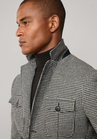 Gray and black houndstooth jacket with a stand-up collar, two front pockets, and brown buttons, worn over a black turtleneck sweater.