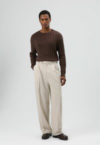 Bruine geribbelde trui, hooggetailleerde crèmekleurige geplooide broek en donkerbruine schoenen, die samen een contrasterend neutraal palet vormen.