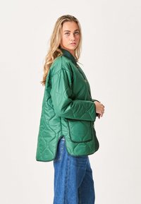 Quiltet grøn jakke med høj krave, trykknapper, sidelommer og løs pasform. Bæres over blå jeans, der fremviser en tekstureret overflade.