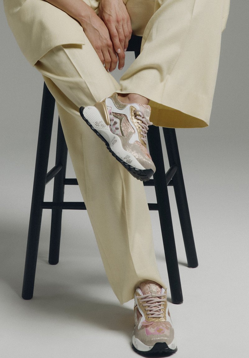 Persona sentada en un taburete negro, vistiendo pantalones anchos crema y zapatillas blancas, rosas y doradas con un patrón, con cordones. Manos descansando sobre la rodilla.