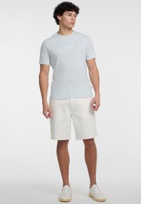 Gaismas zila kokvilnas T-krekls ar īsām piedurknēm, kuram ir neliels balts logo uz krūtīm, apvienots ar baltām šortiem un baltām kedām.