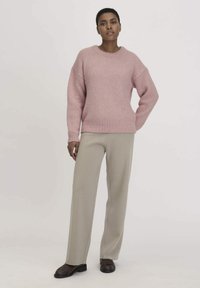 Rosa gestrickter Pullover mit fallenden Schultern, gerippten Bündchen und Saum. Kombiniert mit hellbeigen weitgeschnittenen Hosen und braunen Schuhen.