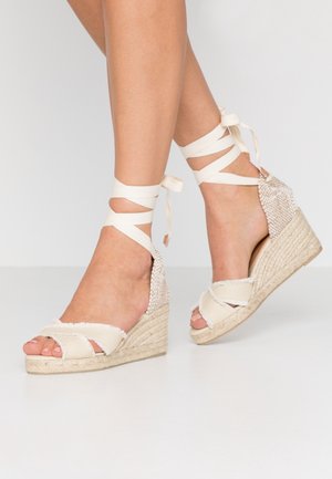 Ben med beige kileespadrilles med krydsede stoffremme og ankelbånd bundet i sløjfer på en lys baggrund.