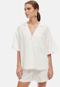 Camicia bianca oversize a maniche corte con bottoni e tasca, realizzata in tessuto leggero, abbinata a pantaloni corti coordinati.