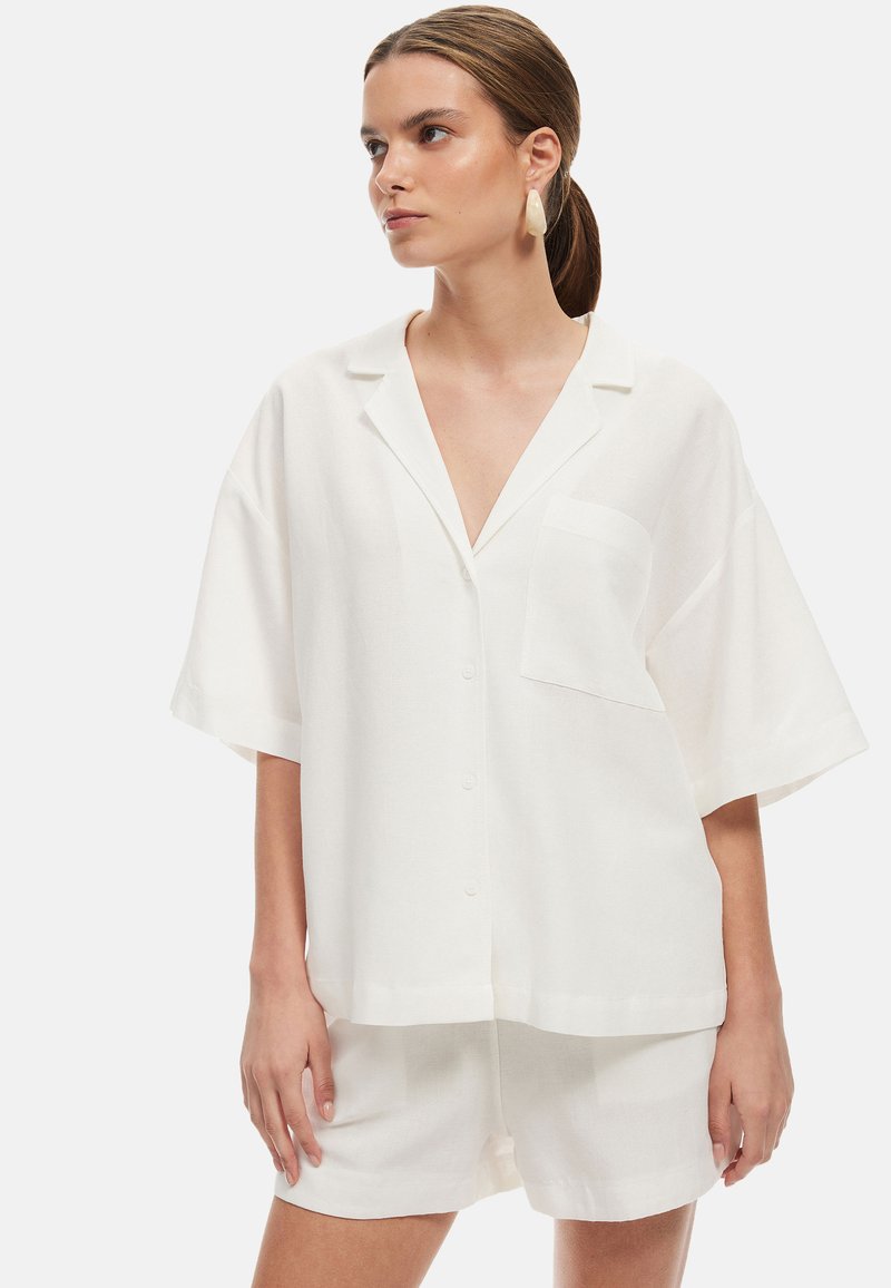 Camicia bianca oversize a maniche corte con bottoni e tasca, realizzata in tessuto leggero, abbinata a pantaloni corti coordinati.