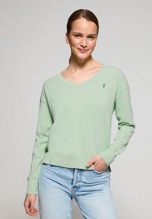 Helles grünes V-Ausschnitt-Objekt, lange Ärmel, gerippter Saum und Bündchen, mit einem kleinen gestickten Logo auf der linken Brust. Getragen mit blauen Jeans.