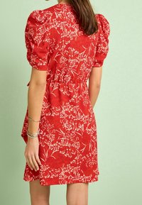 Robe rouge à fleurs avec manches bouffantes, taille ajustée et tissu texturé. Le motif présente des fleurs blanches et des feuilles sur fond rouge.