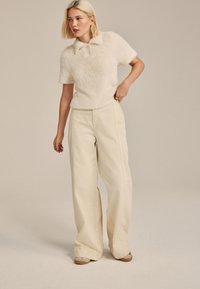 Pull crème duveteux à manches courtes avec col, associé à un pantalon beige taille haute et jambes larges, créant une silhouette décontractée.
