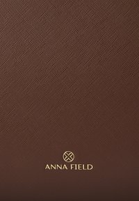 Material marrón texturizado con un ligero brillo, que presenta un logo dorado "ANNA FIELD" y un emblema circular en la parte inferior.