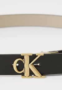 Črn usnjen pas z zaponko "CK" v zlati barvi, ki ima gladko teksturo in kontrastno bež notranjost.