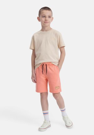 Gutt står med hendene i lommene, iført beige t-skjorte, korallfargede shorts, hvite joggesko og hvite sokker med blå og oransje striper.
