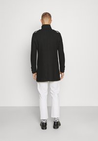 Zwarte lange jas met hoge kraag, gestructureerde schouders en aangepaste pasvorm, gecombineerd met witte broek en zwarte schoenen.