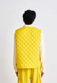 Persona di spalle che indossa un vivace gilet trapuntato giallo, pantaloni gialli abbinati con tasche e una maglietta a maniche lunghe beige su uno sfondo semplice.