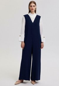 Tumši zils, bez piedurknēm, kombinezons ar dziļu V veida kakla izgriezumu, plašām biksēm un priekšējo rāvējslēdzēju. Saskaņots ar baltu krekliņu ar apkaklīti un bēšām kurpēm.