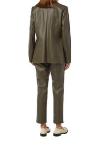 Olivgrüner Kunstledersuit mit taillierter Passform, bestehend aus einem strukturierten Blazer und tapered Hosen, ergänzt durch beige Slipper.