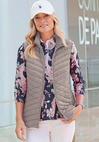 Hellgraue gesteppte Weste über einem blumigen Langarmhemd. Die Weste hat einen Stehkragen und Reißverschlusstaschen. Weiße Mütze mit Logo.