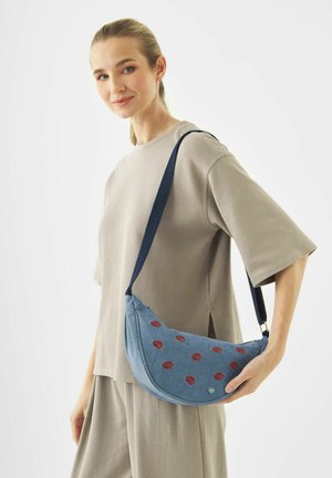 Jonge vrouw in lichtbeige outfit houdt een blauwe schoudertas met rode lippenafdrukken vast tegen een witte achtergrond.