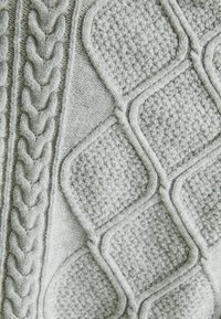 Lichtgrijze gebreide stof met textuurpatronen, waaronder verhoogde diamanten vormen en een kabelbreipatroon langs de linkerrand.