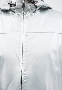 Veste légère argentée avec fermeture éclair à l'avant et capuche ajustable dotée de cordons noirs et roses.