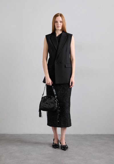 Blazer nero senza maniche con design strutturato, abbinato a una gonna a maglia nera aderente e una piccola borsa nera con frange. Décolleté con punta affilata.