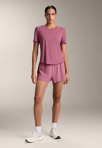 Tričko s krátkým rukávem a kulatým výstřihem a šortky na stažení v měkkém, růžovém materiálu; doplněno o světlé sportovní boty a bílé ponožky do kotníků.