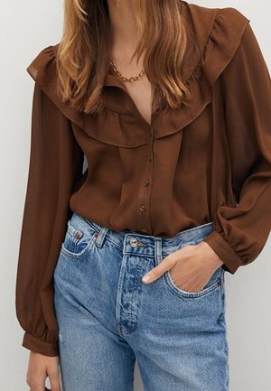 Kvinde iført en brun bluse med flæser, stoppet ned i højtaljede lyseblå jeans, med den ene hånd i lommen og et guldkædehalskæde.