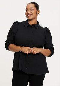 Zwarte blouse met knopen en pofmouwen, gemaakt van zachte stof en met een gebogen zoom. Model draagt een gouden armband en heeft een neutrale achtergrond.