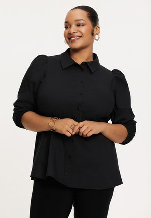Zwarte blouse met knopen en pofmouwen, gemaakt van zachte stof en met een gebogen zoom. Model draagt een gouden armband en heeft een neutrale achtergrond.