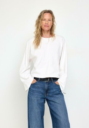 Donna con capelli biondi mossi che indossa una maglia bianca a maniche lunghe e jeans blu, in piedi con le mani nelle tasche davanti a uno sfondo semplice.