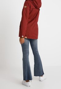 Giacca impermeabile rossa con cappuccio, caratterizzata da toppe e dettagli con bottoni, abbinata a jeans blu svasati e usurati e sneakers bianche.