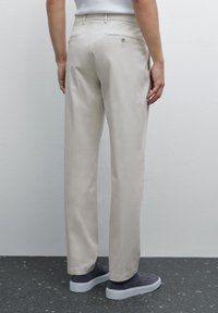 Light beige trousers with straight cut, back pockets, and belt loops. Worn with grey slip-on shoes, displayed from the back.