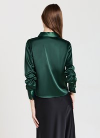 Woman with long brown hair wearing a dark green satin blouse and black silky skirt, shown from the back against a plain background.