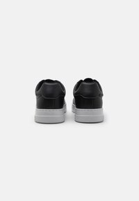 Adidași negri din piele cu o textură netedă, talpă din cauciuc alb și un design minimalist. Prezintă un logo pe tocul pantofului.