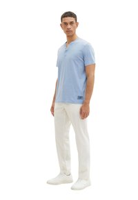 Chemise henley en coton bleu clair à manches courtes, avec une patte de boutonnage à deux boutons et un détail en étiquette noire, associée à un pantalon blanc et des baskets blanches.