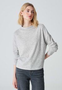Hellgrauer Pullover mit strukturiertem Strickmuster, 3/4-Ärmeln und hohem Kragen. Kombiniert mit dunklen Jeans.