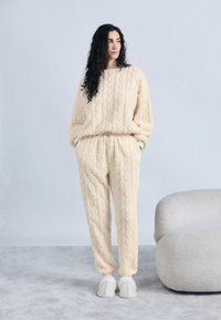 Γυναίκα με μακριά σκούρα μαλλιά, φορούσα κρεμ υφασμάτινο ρουχισμό για χαλάρωση και παντόφλες, στέκεται δίπλα σε μια στρογγυλεμένη λευκή ταπετσαρισμένη καρέκλα πάνω σε γκρι χαλί.